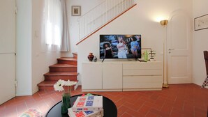 A living room with a staircase, a television on a white cabinet, a vase with flowers, and a coffee table with books. - Marsili Suite (Florence)