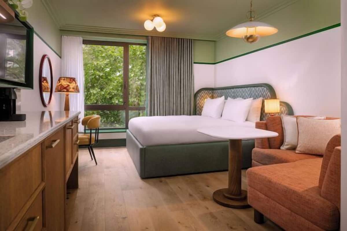 A hotel room with a bed, a sofa, a desk with a chair, a TV, and a window with curtains.