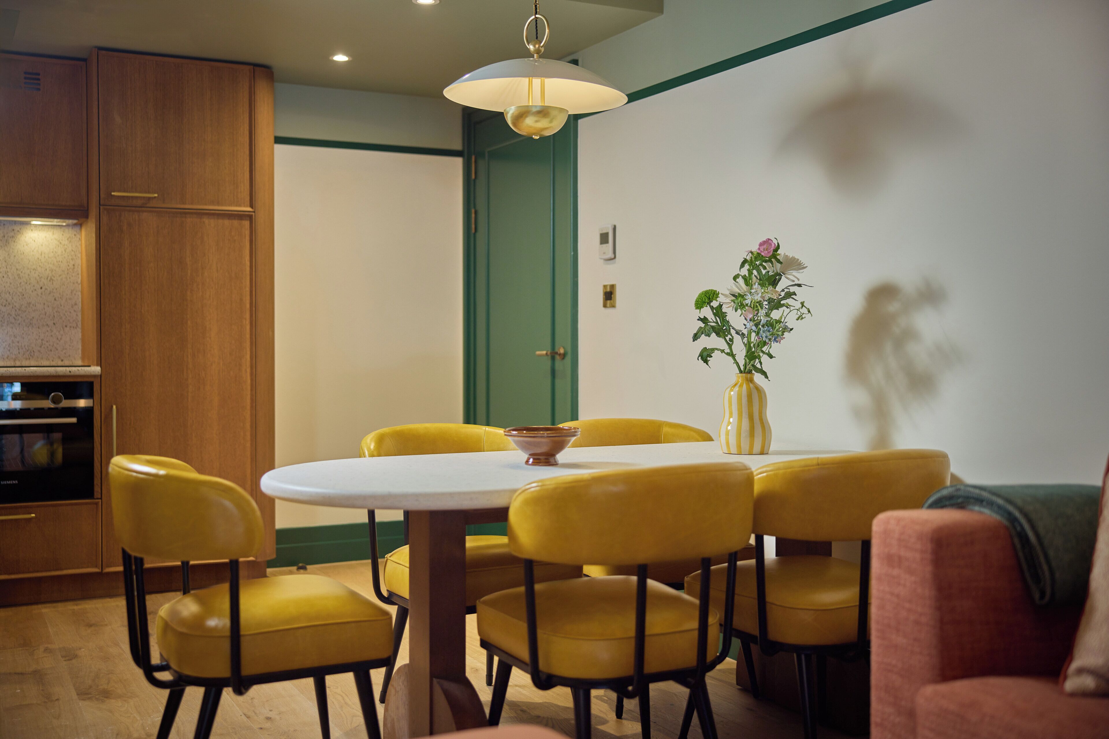 A dining area with a round table, yellow chairs, a vase with flowers, and a green door in the background.