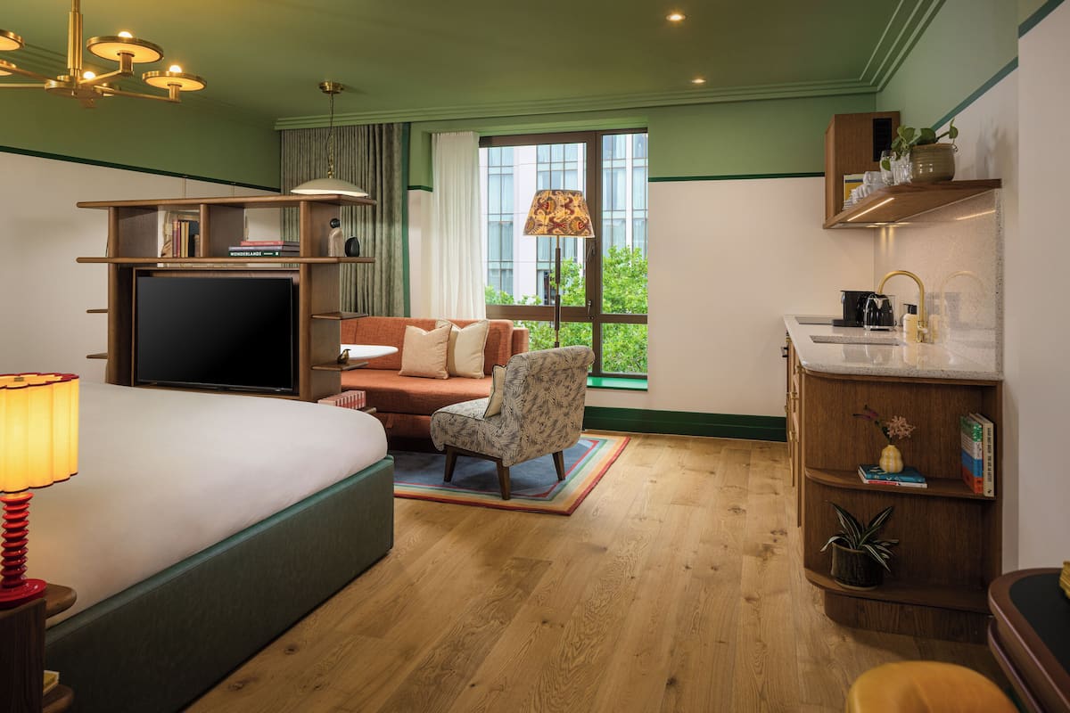 A hotel room with a bed, a sofa, a chair, a TV, a desk, a lamp, a window with a view, and a sink.