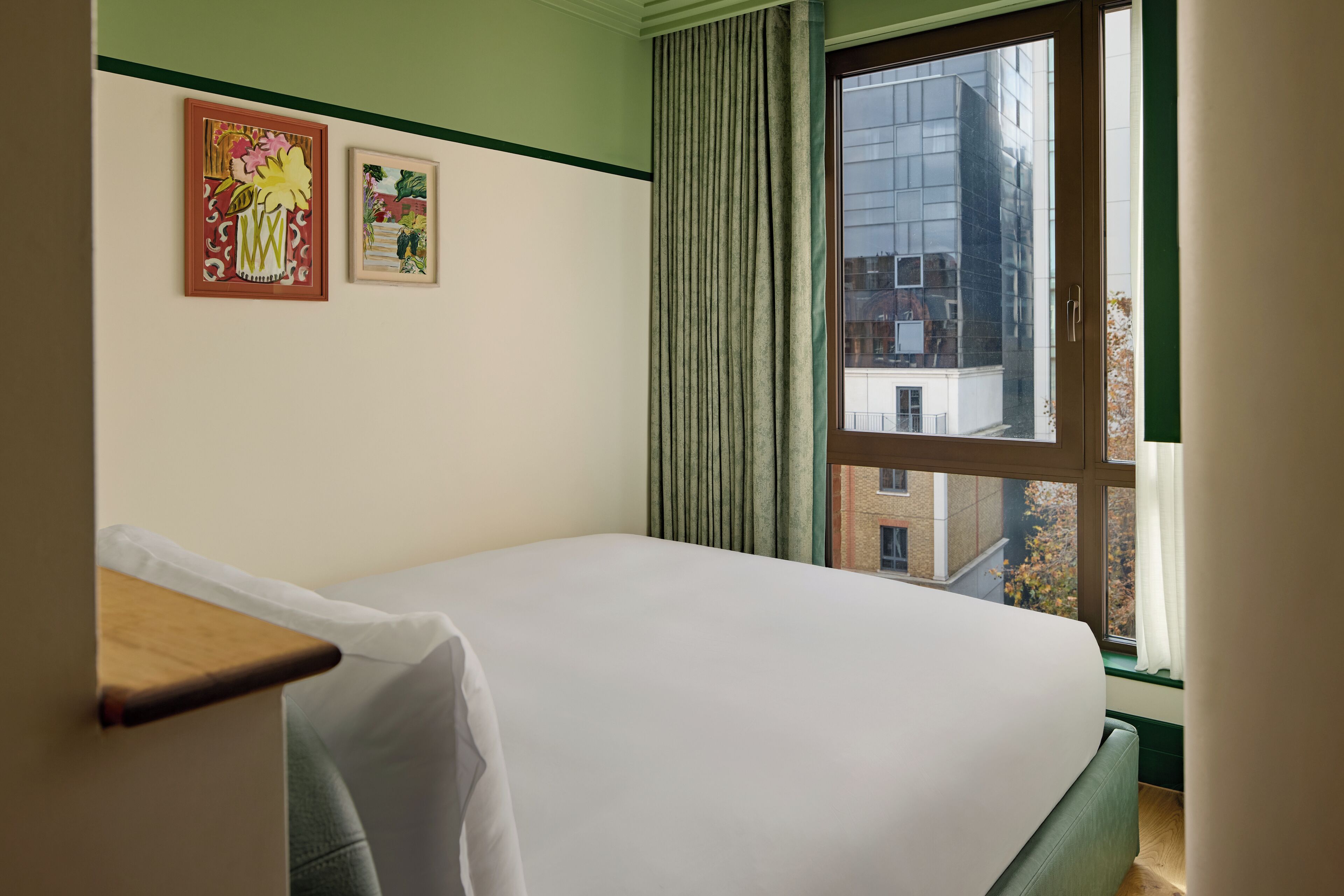 Ein Hotelzimmer mit einem Bett, einem Fenster mit Vorhängen und zwei gerahmten Kunstwerken an der Wand.