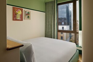 Ein Hotelzimmer mit einem Bett, einem Fenster mit Vorhängen und zwei gerahmten Kunstwerken an der Wand.