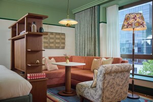 Ein gemütliches Zimmer mit Holzregal, einem runden Tisch, einem gemusterten Sessel und einer Stehlampe.
