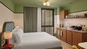 A hotel room with a bed, a nightstand, a lamp, a window with curtains, a kitchenette with cabinets, and a dining area with a chair. - The July London Victoria (London)