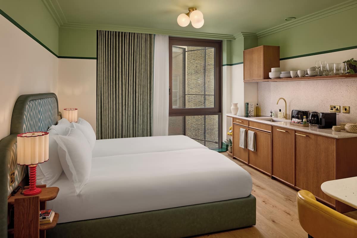 A hotel room with a bed, a nightstand, a lamp, a window with curtains, a kitchenette with cabinets, and a dining area with a chair.