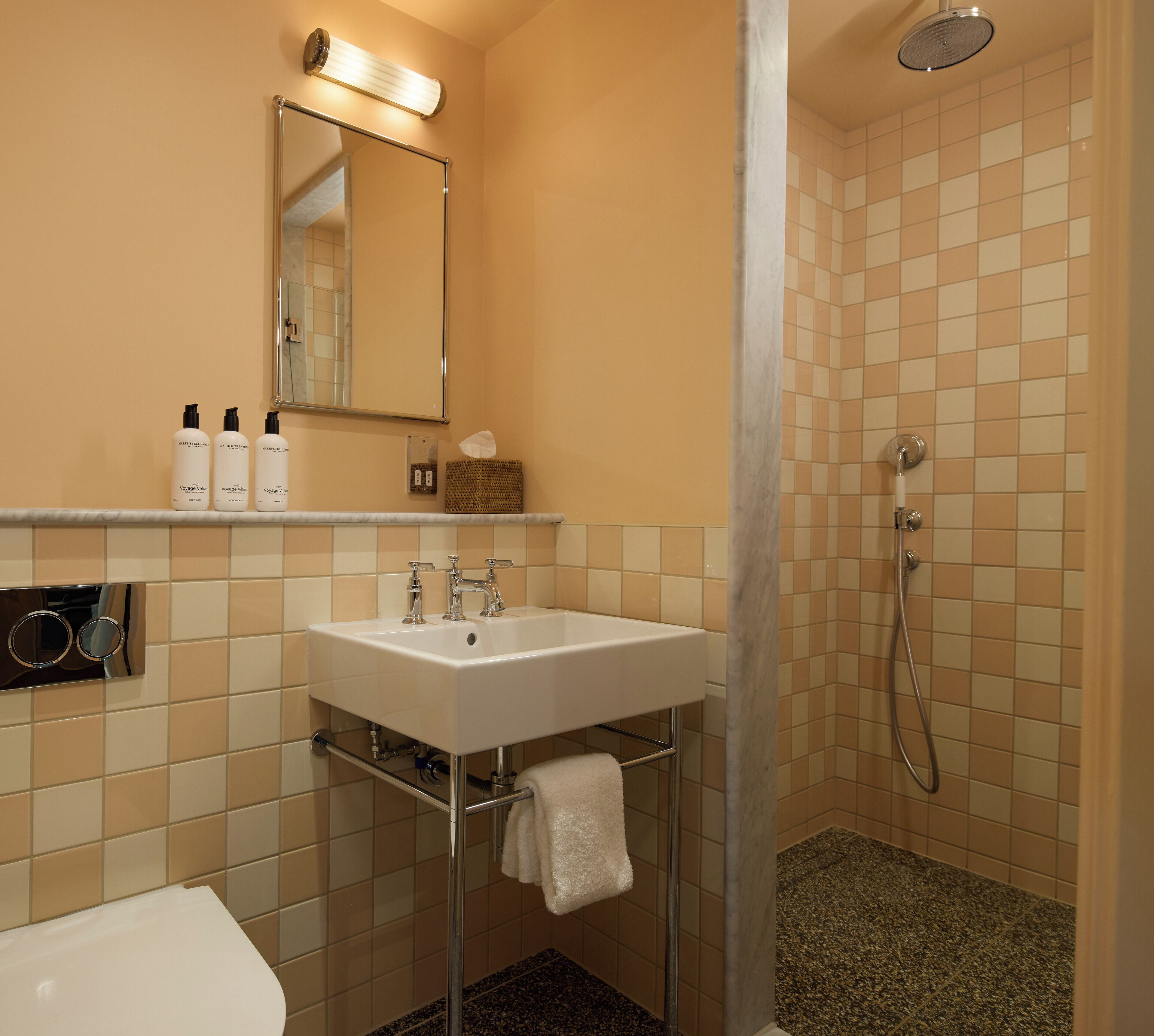 Een badkamer met een wit wastafel, spiegel en douche. Er liggen toiletartikelen op het aanrecht en een handdoek hangt aan de muur.