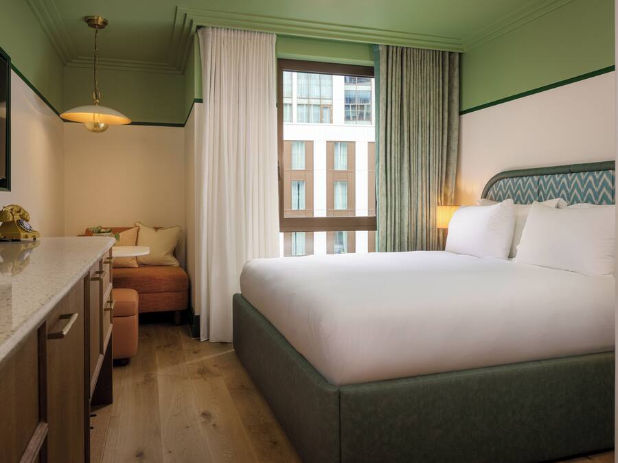 Quarto de hotel com cama, secretária, cadeira e janela com cortinas.