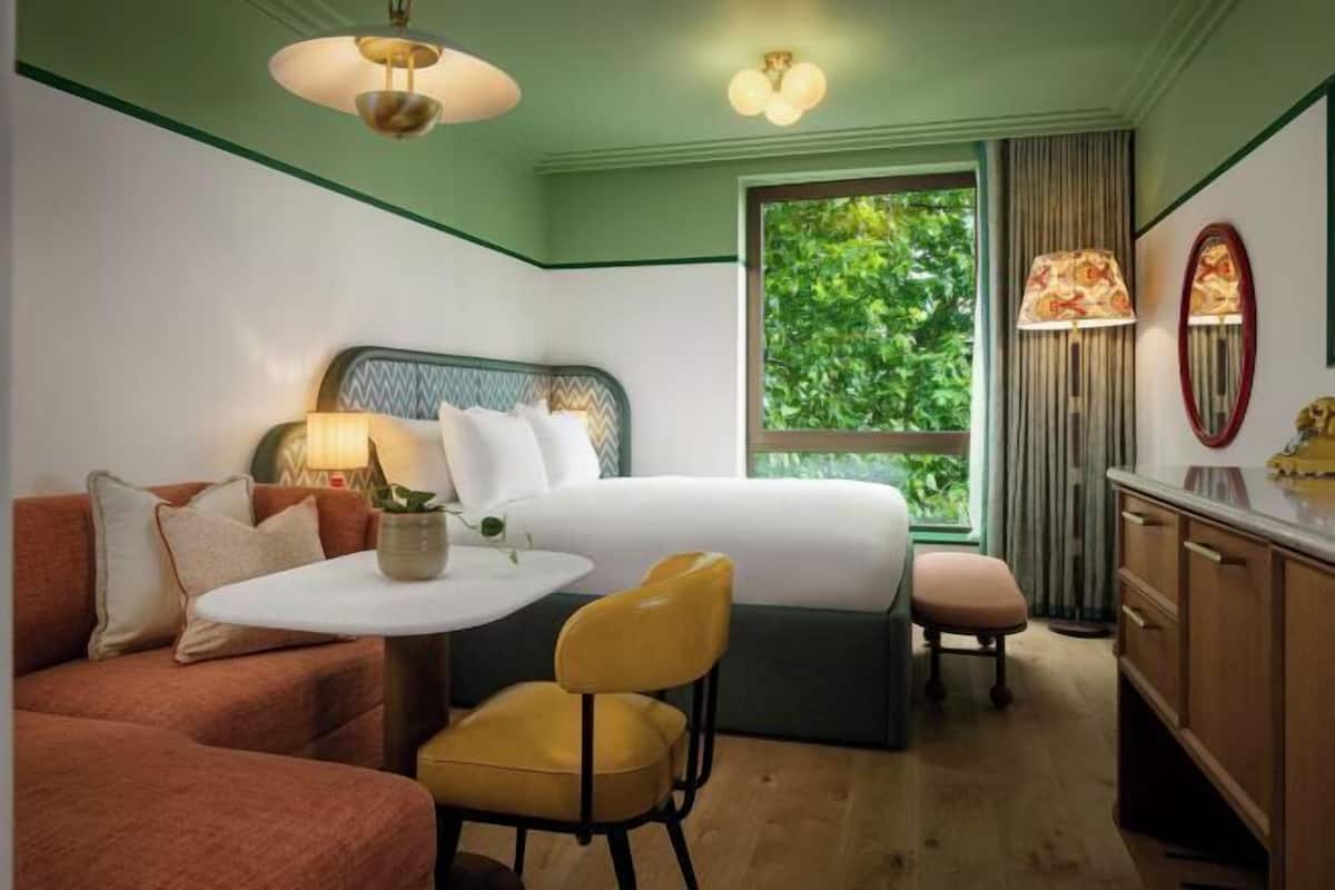 A hotel room with a bed, a sofa, a desk, a chair, a lamp, a mirror, and a window with a view of greenery.