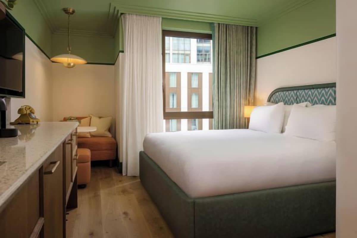 A hotel room with a bed, a desk, a chair, a window with curtains, and a lamp.