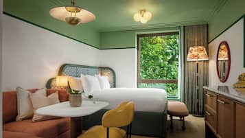 A hotel room with a bed, a sofa, a chair, a small table, a desk, and a window with a view of greenery.