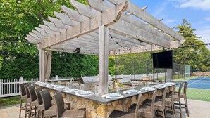 Un espace bar extérieur doté d’un comptoir en pierre, de chaises en osier, d’une pergola et d’un écran de télévision.