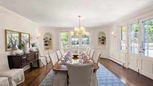 Une salle à manger avec une longue table dressée pour six personnes, des murs blancs et de grandes fenêtres.
