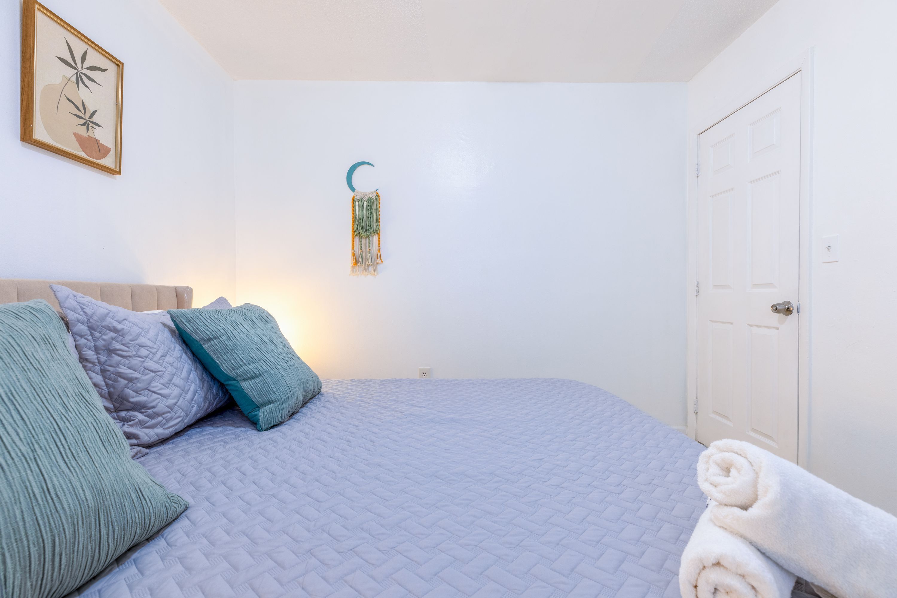 A neatly made bed with a quilted cover, a wall-mounted crescent moon decoration, and a rolled towel on the floor.