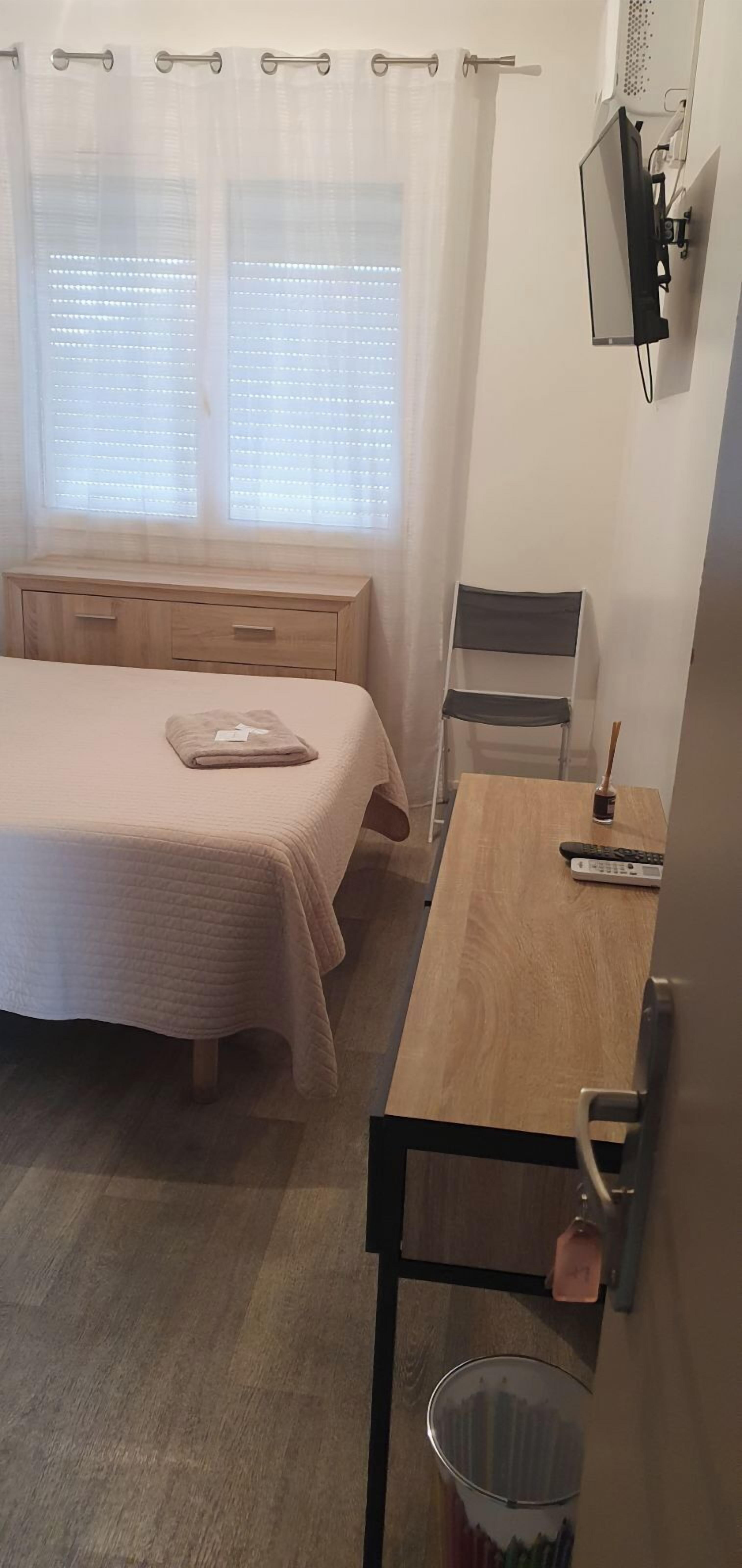 Ein Schlafzimmer mit Bett, einem Holztisch, einem Stuhl, einem Fernseher und einem Fenster mit Vorhängen.