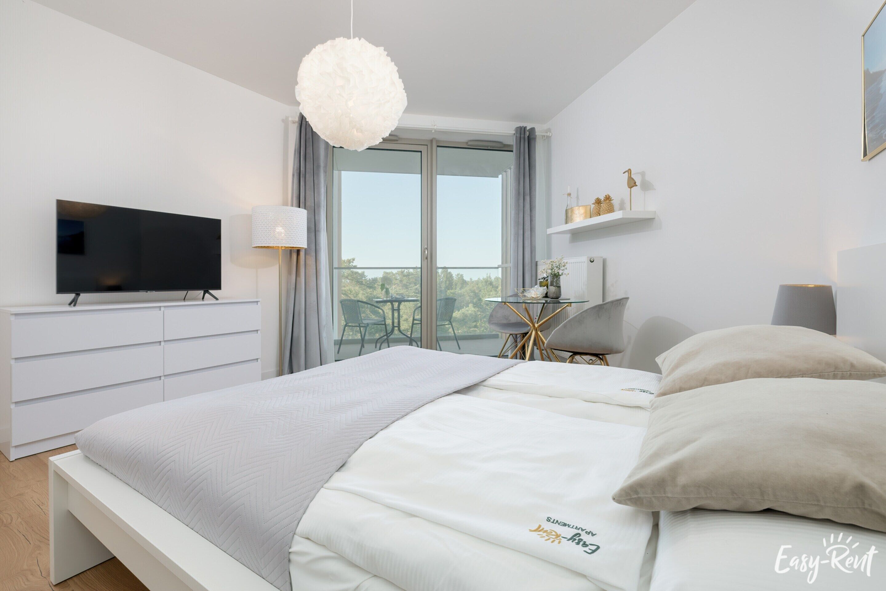 A modern bedroom with a large bed, a flat-screen TV, a balcony with a table and chairs, and a view of the outdoors.