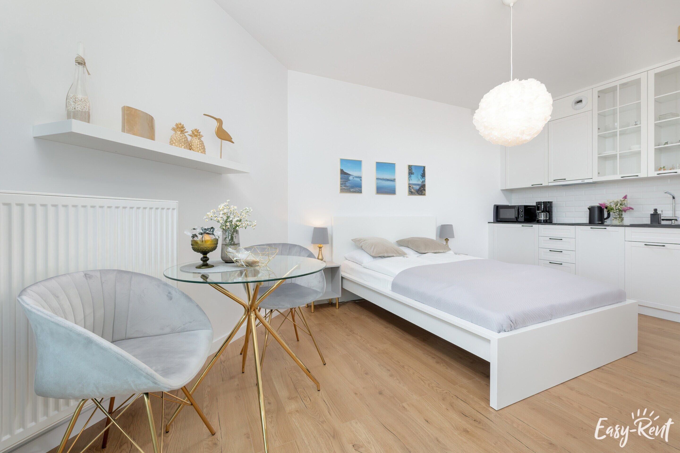 Une chambre moderne avec un lit blanc, une table en verre et une chaise. On y trouve également un coin cuisine avec des meubles blancs et un four à micro-ondes.