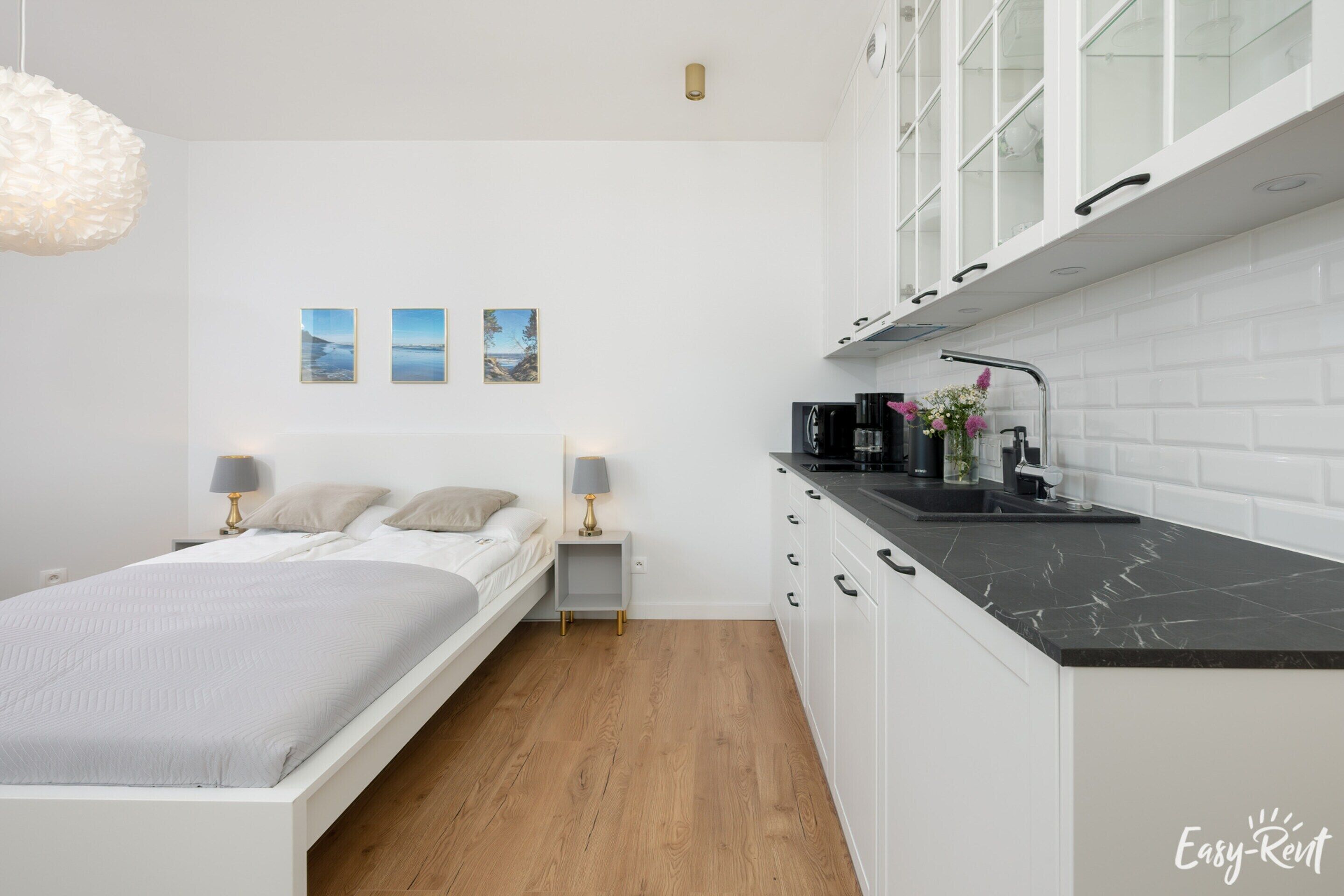 A modern bedroom with a bed, bedside tables, and a small nightstand. Adjacent to the bedroom is a compact kitchenette with a black countertop, white cabinets, and a sink.