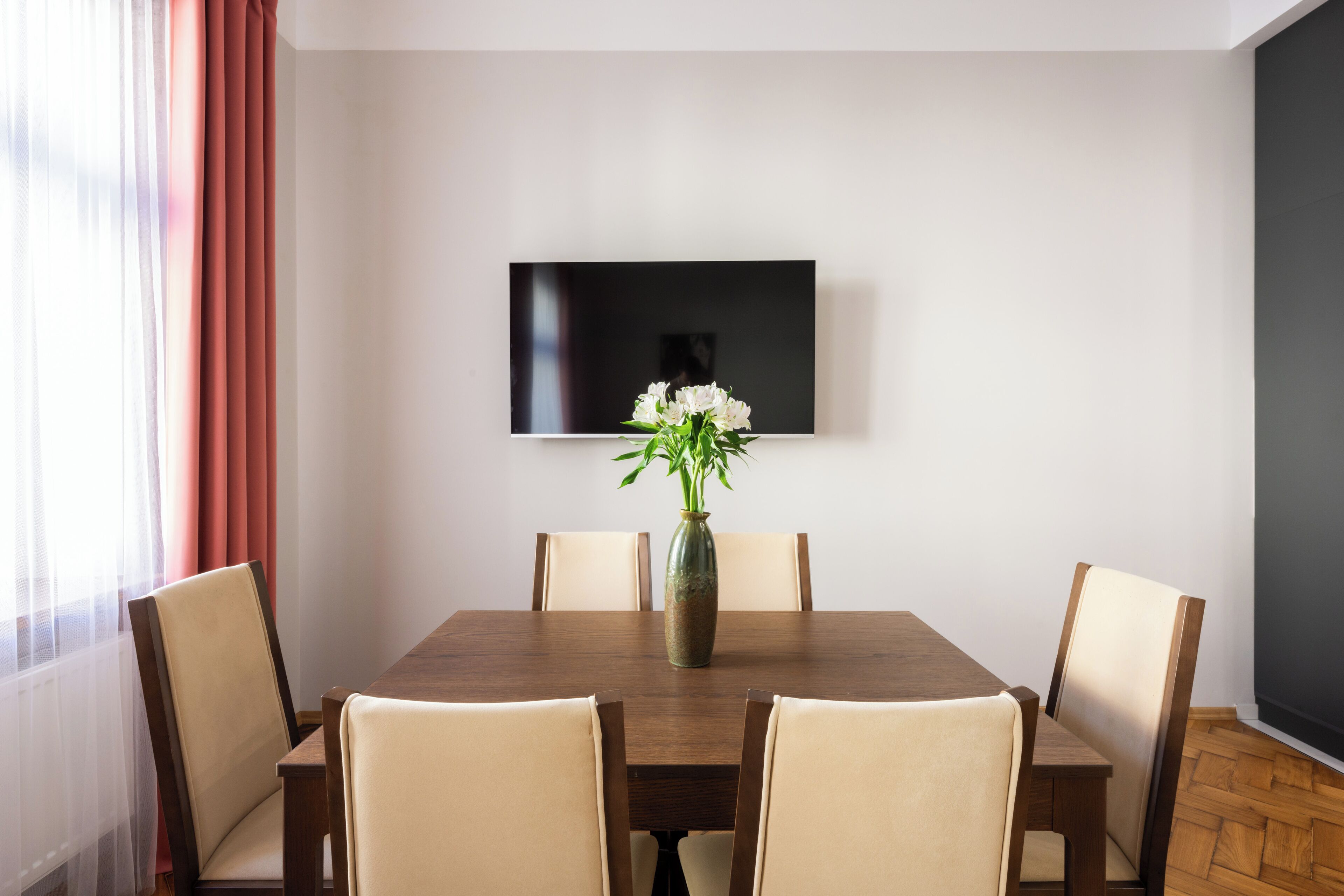 Een eethoek met een houten tafel, zes stoelen, een vaas met bloemen en een flatscreen televisie aan de muur.