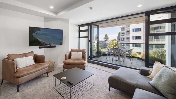 A modern living room with leather sofas, a coffee table, and a large window offering a view of a balcony and neighboring buildings. - Ocean Eleven Mt Maunganui (Mount Maunganui)