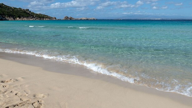Una playa con agua turquesa cristalina, orilla de arena y colinas rocosas al fondo.