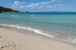 Uma praia com águas turquesa cristalinas, areal arenoso e colinas rochosas ao fundo.