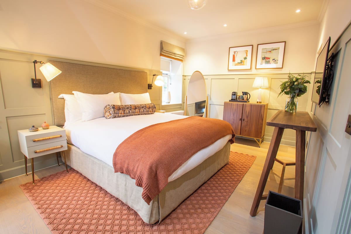 A hotel room with a bed, bedside tables, a nightstand, a desk, a chair, a lamp, a mirror, and framed artwork on the wall.