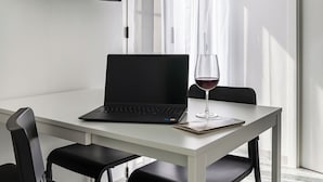 Un portátil sobre una mesa blanca con una copa de vino tinto, sillas negras y un televisor montado en la pared.