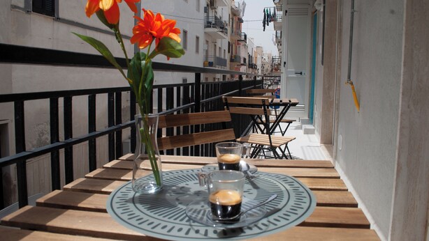Un balcón con una mesa lista para dos personas, con un jarrón de flores y dos tazas de café.