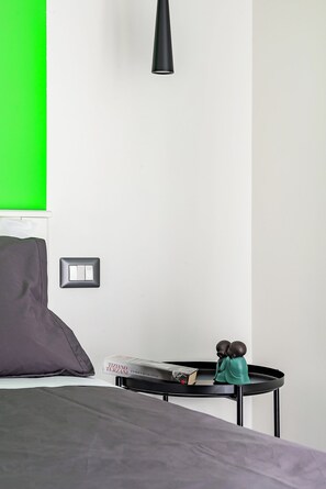 A bed with a grey blanket, a black bedside table with a book and a figurine, a green wall, and a black pendant light.
