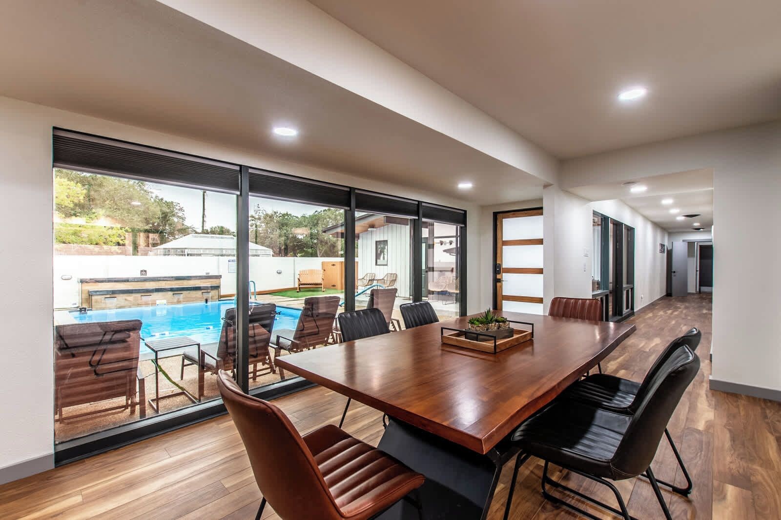 A spacious indoor area with a large wooden table and chairs, large windows offering a view of an outdoor swimming pool.