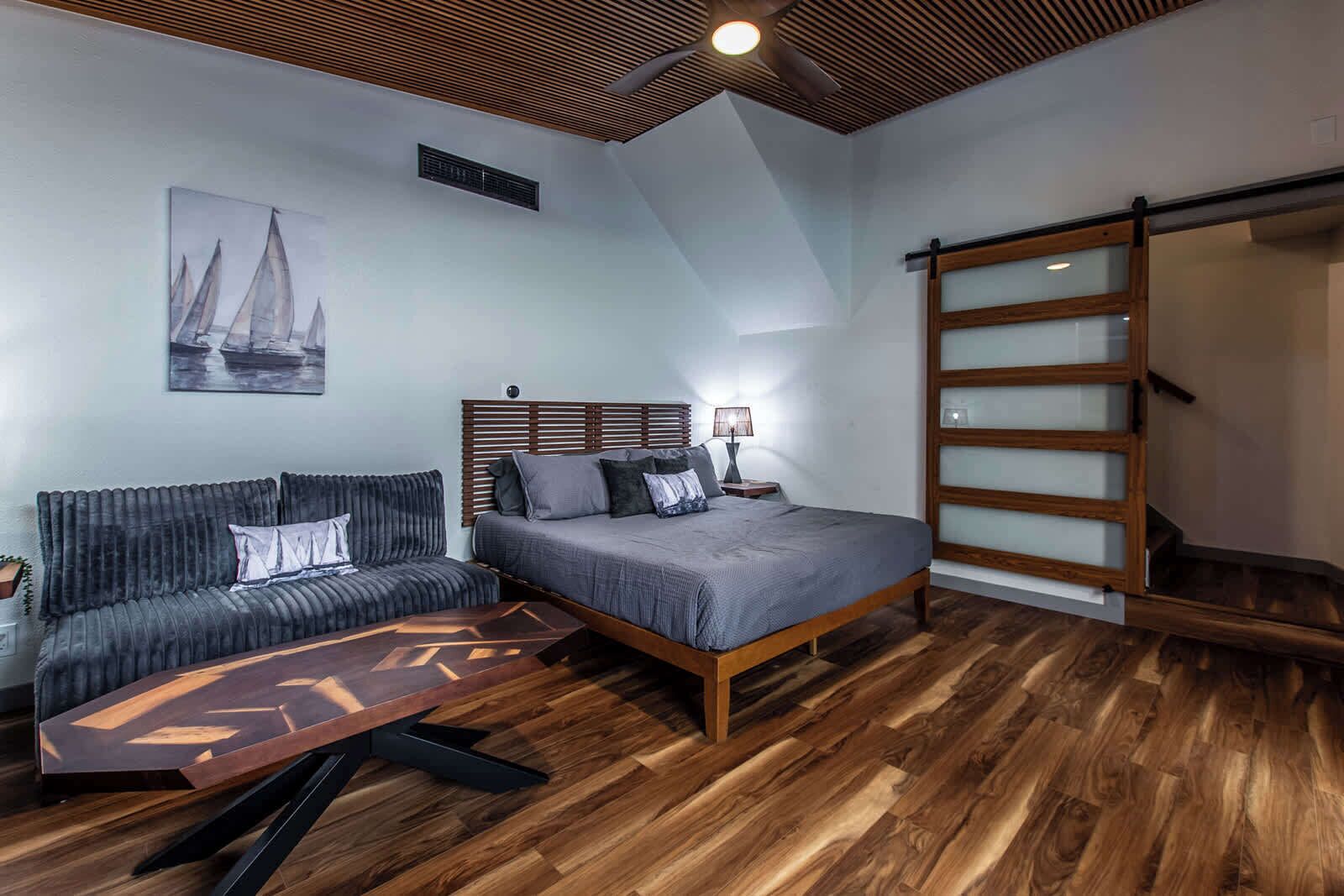 Ein modernes Schlafzimmer mit Holzboden, einem Bett mit grauer Bettdecke, einem dunkelgrauen Sofa und einem Holztisch.