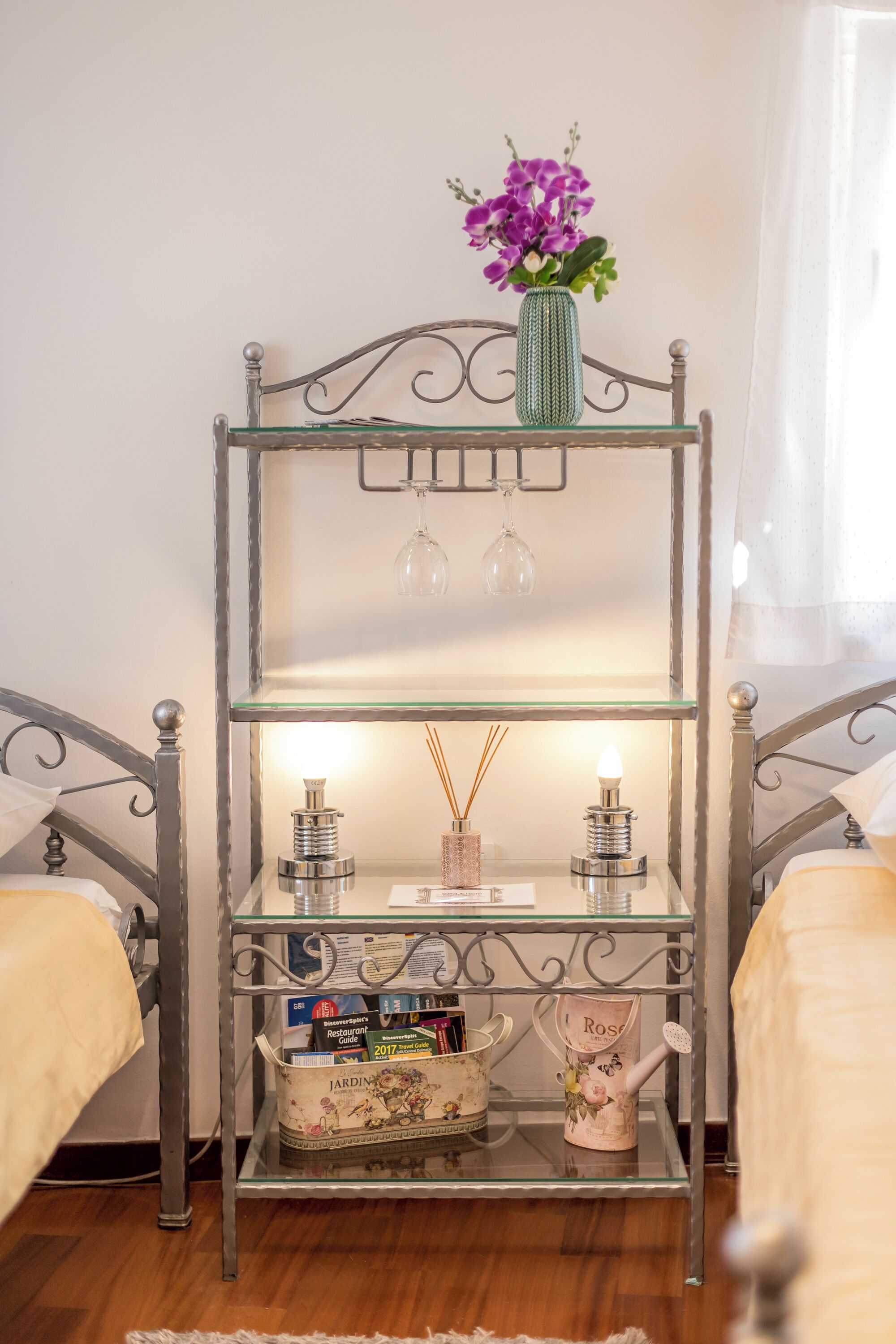 A metal-framed shelf unit with a vase of purple flowers, a glass candle holder, and a watering can on the top shelf. The middle shelf holds a small lamp, a candle, and a decorative item. The bottom shelf has a box labeled "JARDIN" and a watering can.