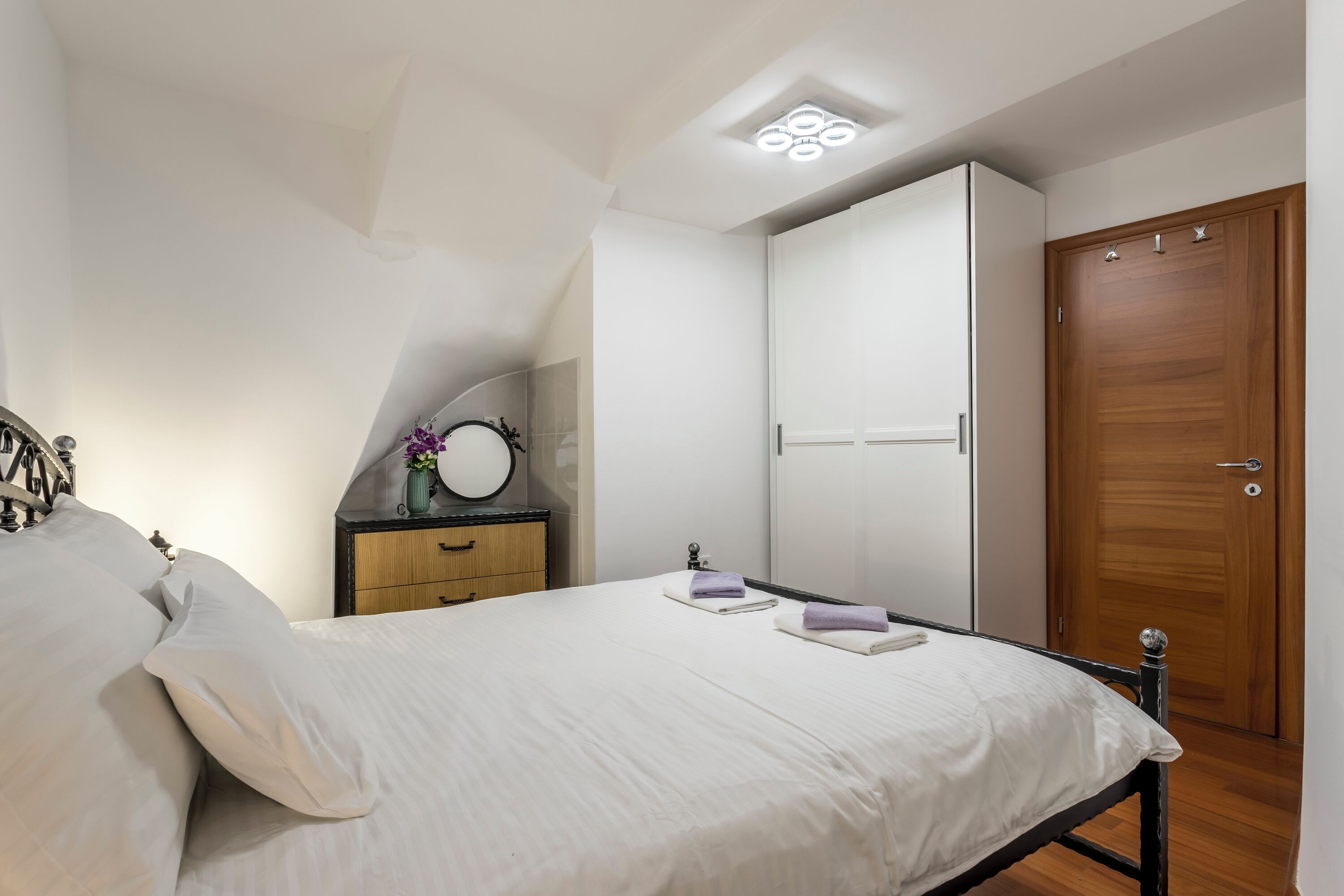 A bedroom with a bed, a wooden wardrobe, a small bedside table with a vase, and a ceiling light fixture.
