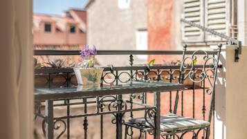 Un balcón con una mesa y una silla de metal, una planta en maceta con flores moradas, y vistas de edificios al fondo.