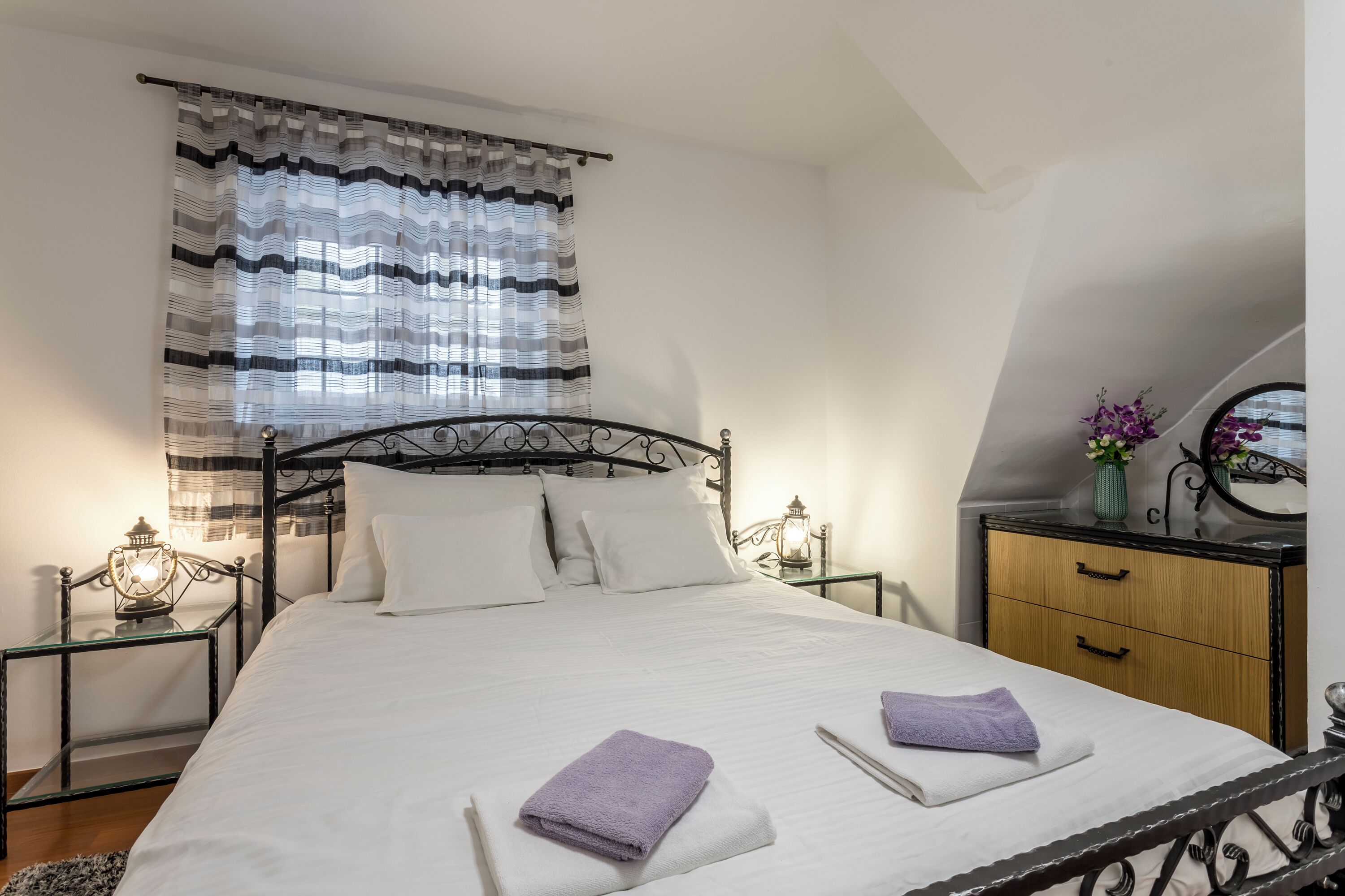 A neatly made bed with white linens and two folded purple towels, a bedside table with a lamp, a small wooden nightstand, a mirror, and a window with striped curtains.