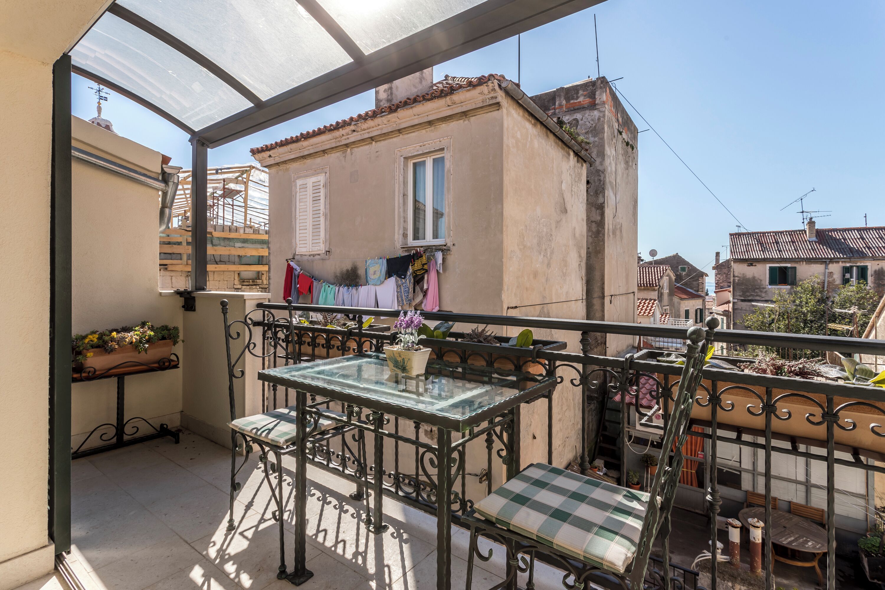 A balcony with a glass table, metal chairs, and a checkered cushion. There is a clothesline with laundry and a potted plant on the table.