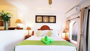 Una cama bien hecha con una toalla verde, un cabecero de madera y una colcha blanca. Encima de la cama, tres cuadros enmarcados; una ventana con cortinas y un aire acondicionado de pared.