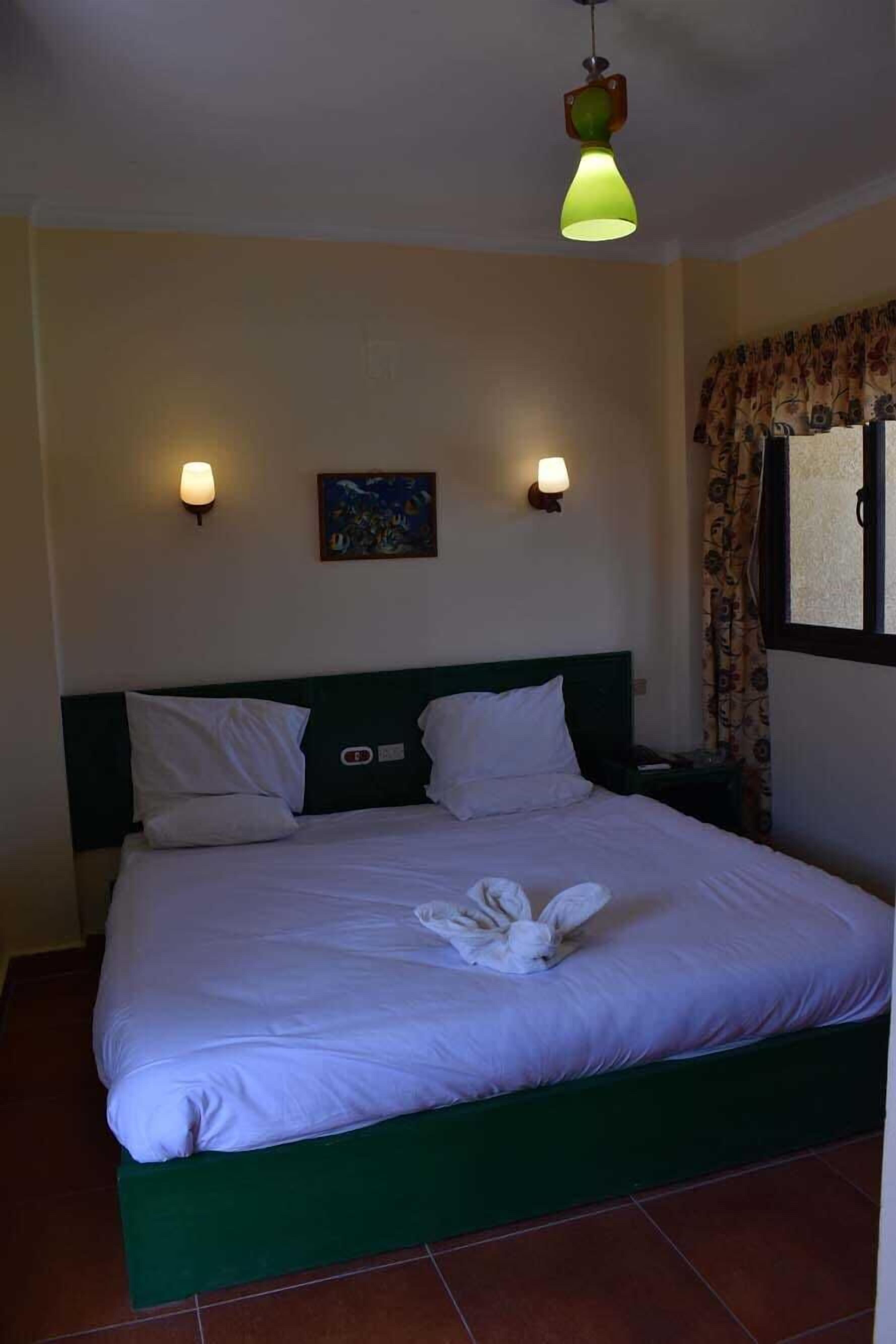 Un lit bien fait, avec du linge de lit blanc et une tête de lit verte, deux lampes fixées au mur et un tableau au-dessus du lit.