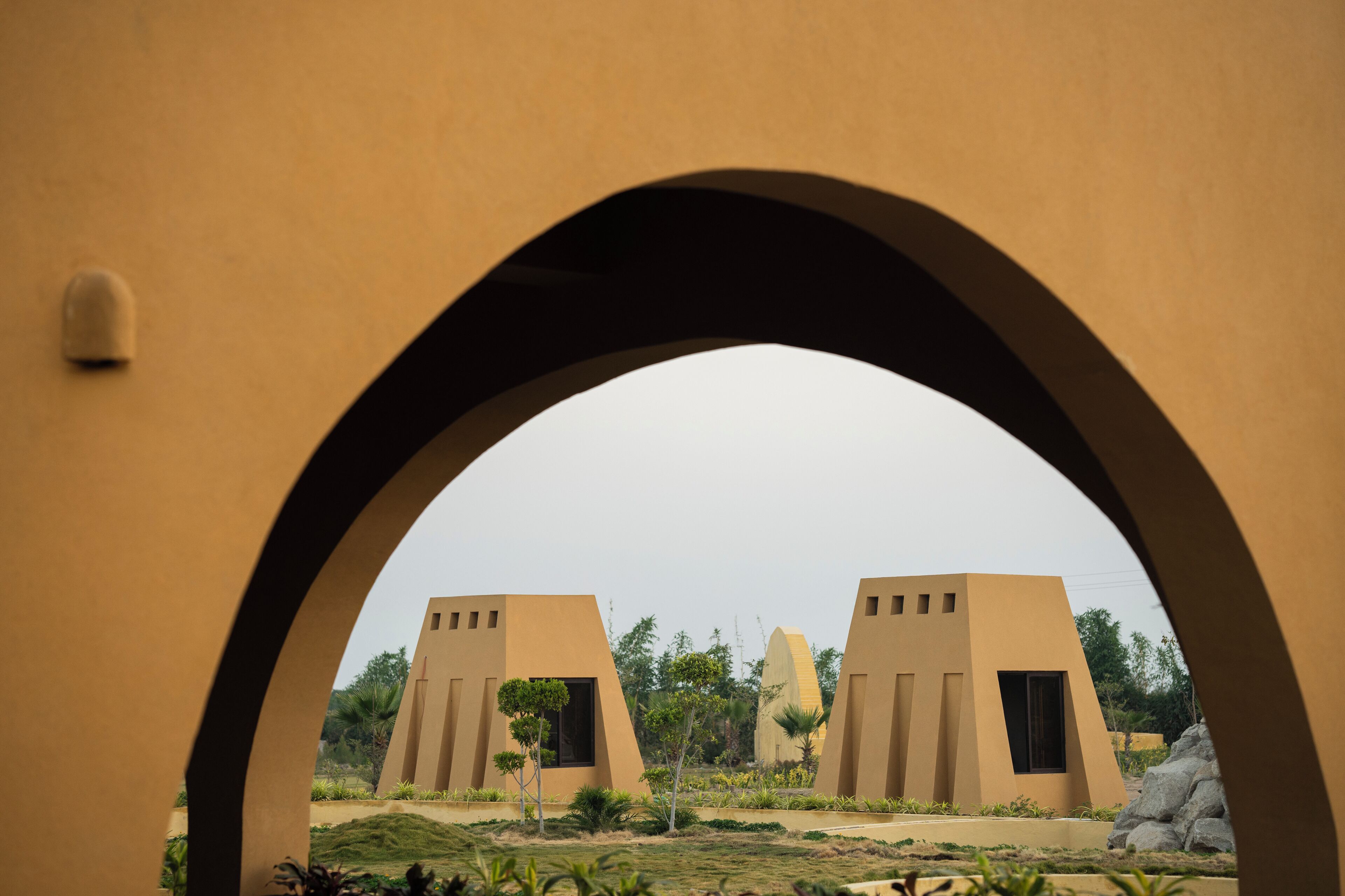 A view through an archway of two beige buildings with modern architecture, surrounded by greenery.