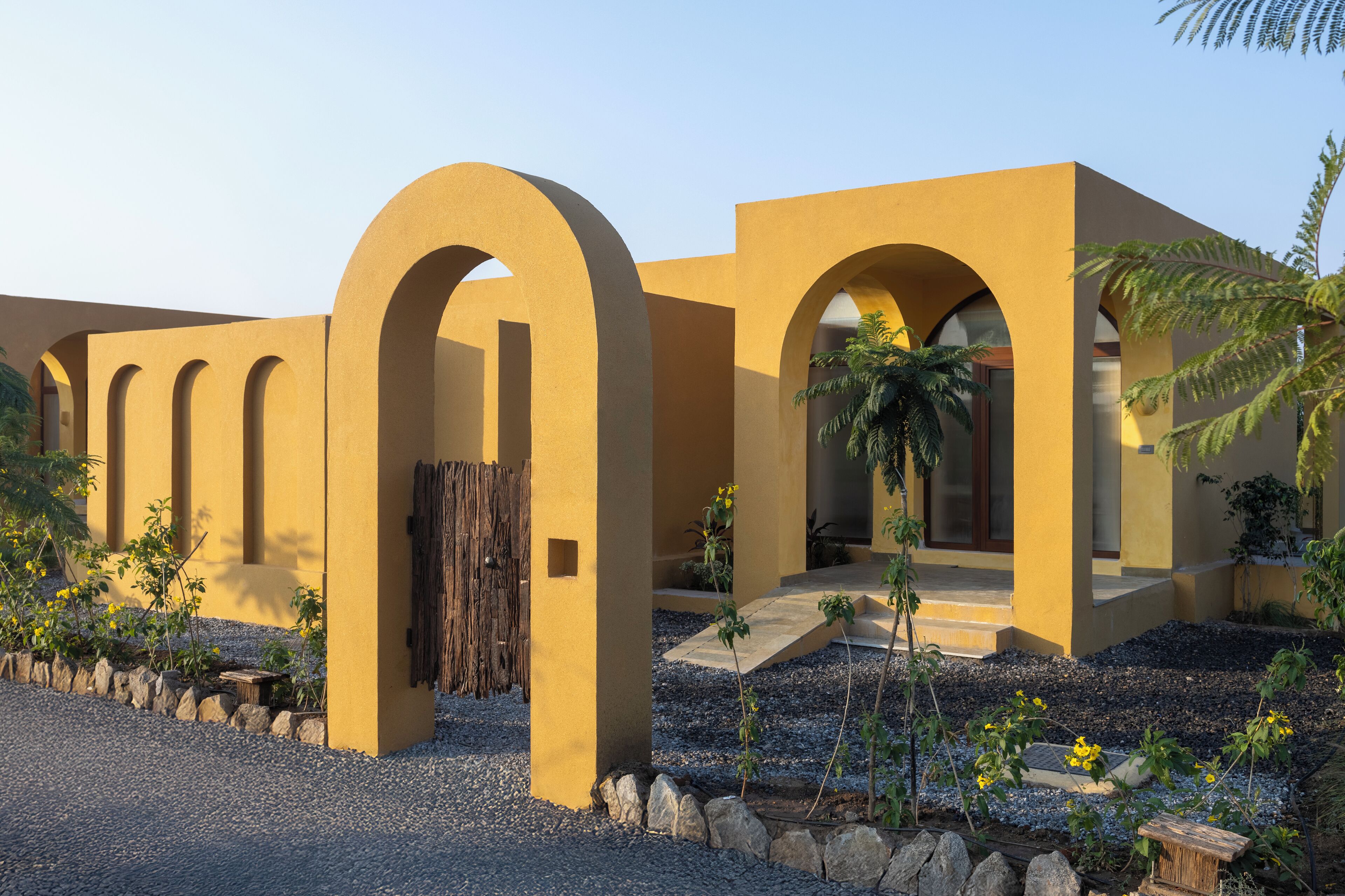 A building with yellow walls and multiple arches, featuring a gravel pathway and small plants.