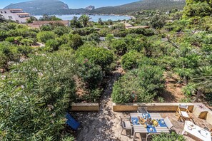 Um pátio com jardim, mesa posta para refeições, cercado por vegetação exuberante e vista para o mar.