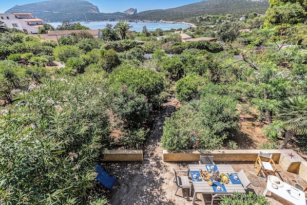 Un patio jardín con mesa lista para comer, rodeado de exuberante vegetación y vistas al mar.