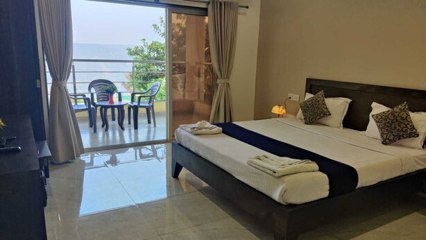 A hotel room with a large bed, a balcony with chairs, and a view of the sea. - Heaven Hills Resort (Mahabaleshwar)