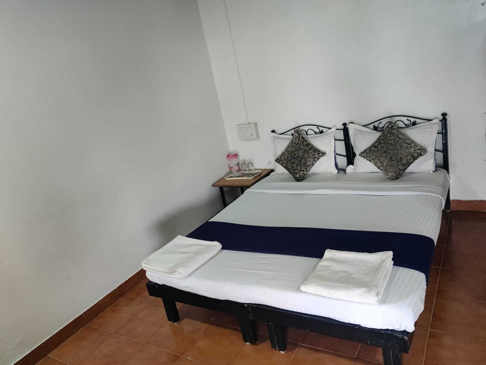 A neatly made bed with white linens and two decorative pillows, a small bedside table with a lamp, and a wall socket.