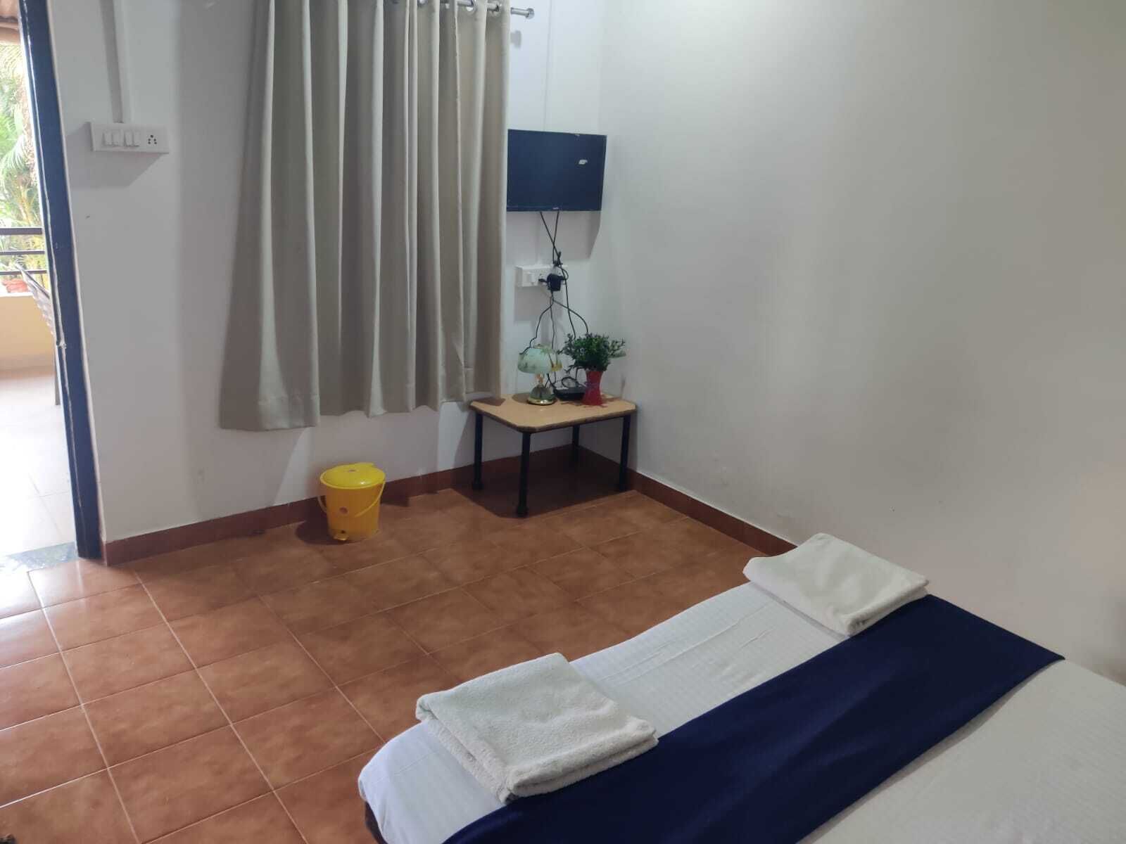 A room with a bed, a small table with a lamp and a plant, a yellow bucket, and a TV mounted on the wall.