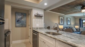 A modern kitchen with a marble countertop, built-in dishwasher, and a large sink. There is a dining area with a blue sofa and a ceiling fan.