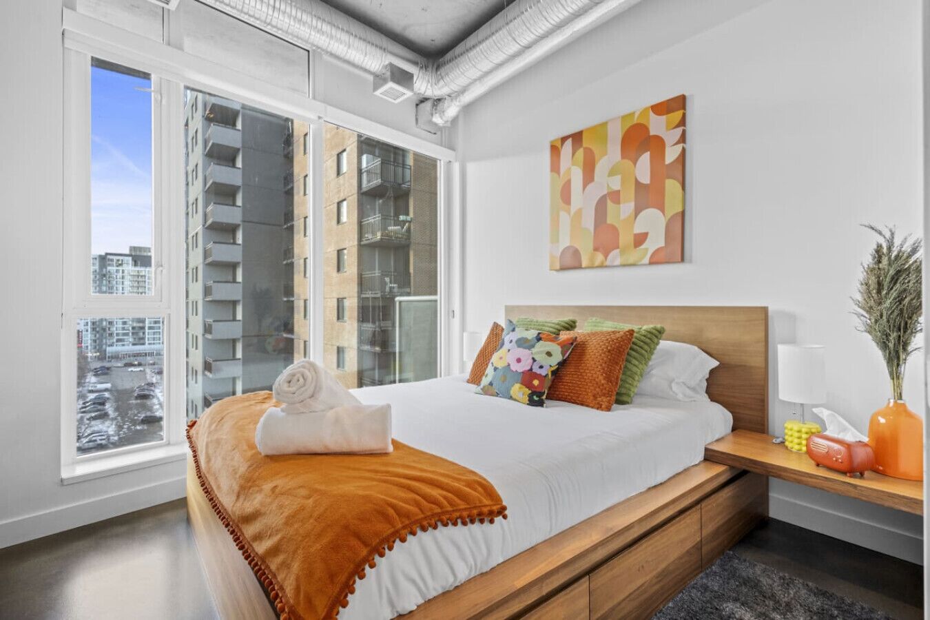 Ein modernes Hotelzimmer mit einem großen Bett, einem Holzkopfstück, Blick auf die Stadt durch raumhohe Fenster und einem farbenfrohen abstrakten Gemälde an der Wand.