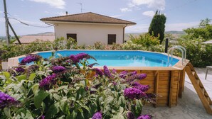 Un bain à remous en bois avec une piscine à l’eau d’un bleu limpide, entouré d’une végétation luxuriante et de fleurs violettes, avec une maison en arrière-plan.