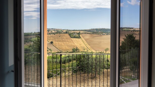Une vue depuis un balcon doté d’une rambarde en métal, donnant sur un paysage rural avec des champs et des bâtiments situés au loin.