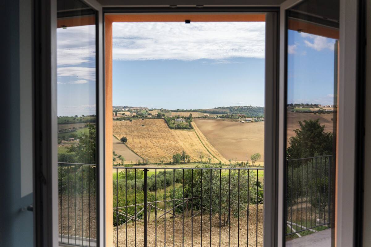 A view from a balcony with a metal railing, overlooking a rural landscape with fields and distant buildings.